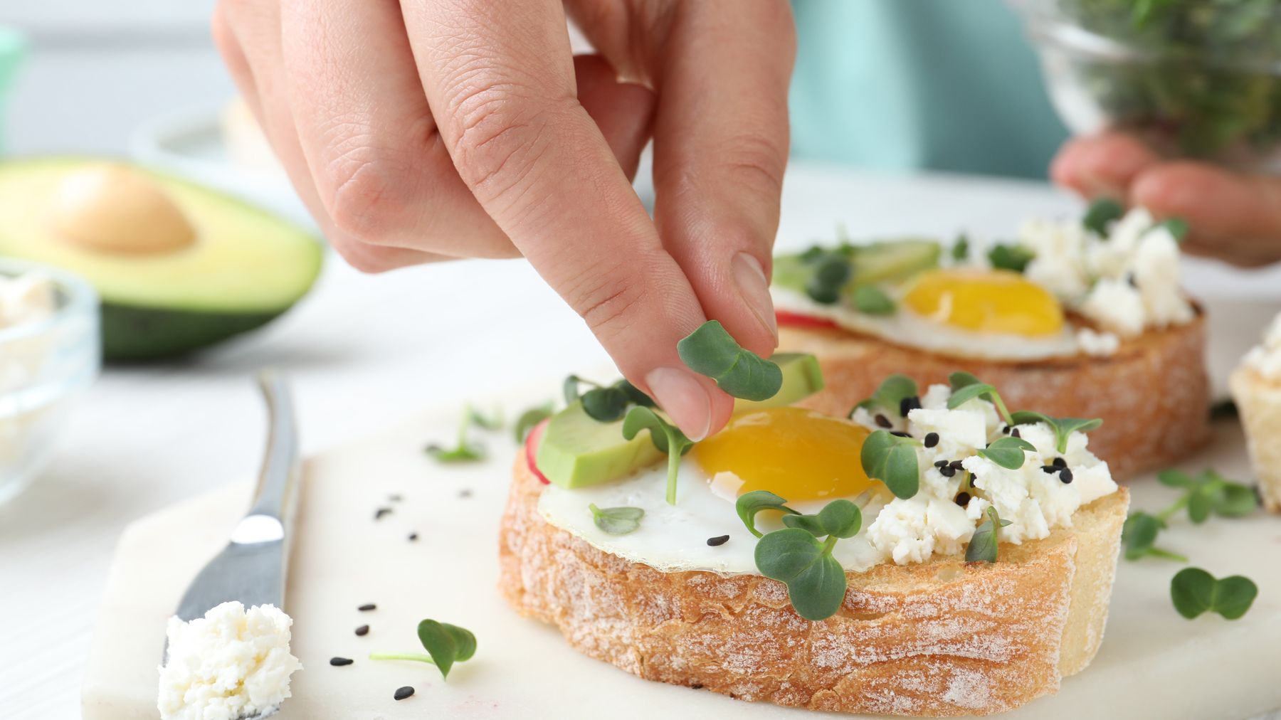 A hand adding microgreens to toast topped with a fried egg, avocado and feta